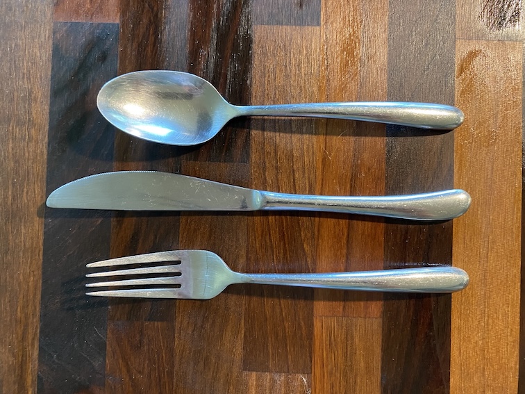 A spoon, knife, and fork are horizontally lined up on a dark wood countertop.