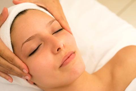 Close-up of a woman's face receiving a facial massage. Her eyes are closed while two hands rub her temples.