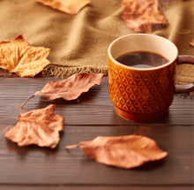 A warm cup of cider in a tan embossed mug rests on a table surrounded by autumn leaves.