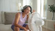 A middle-aged woman in a tank top and shorts sits in front of a white fan, trying to cool down.